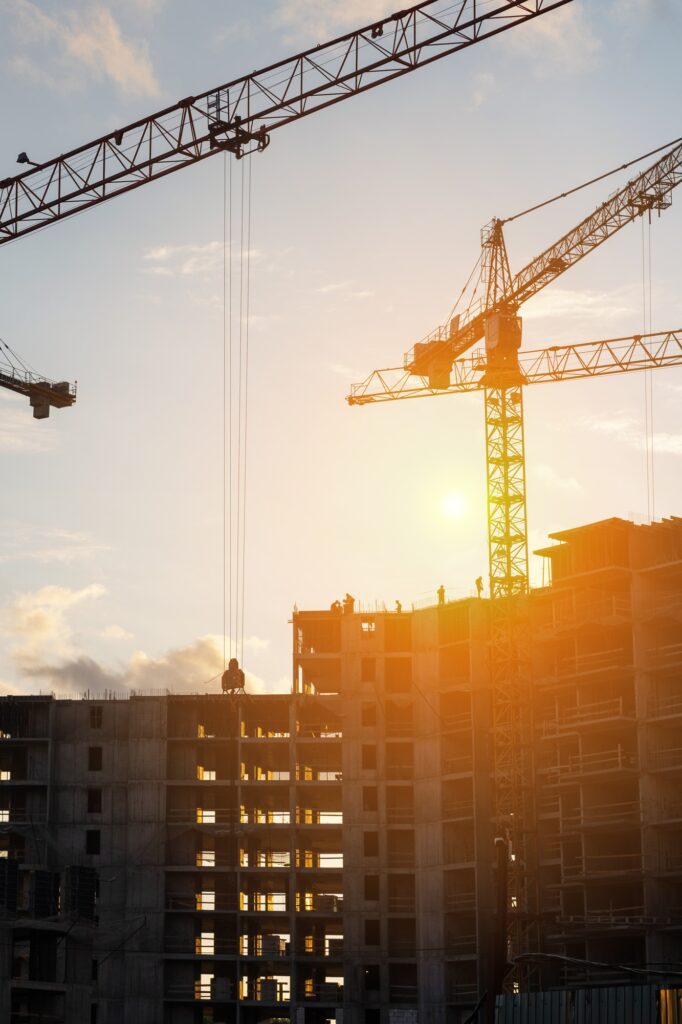Cranes operate at a construction site during sunset, with workers actively engaged in building